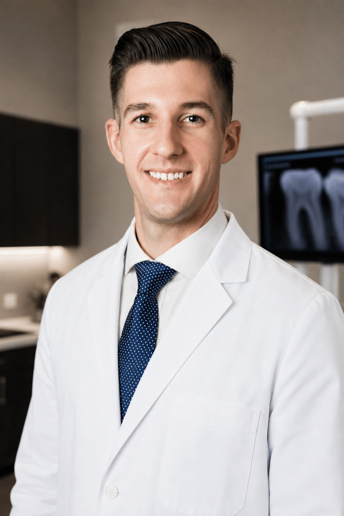 A smiling man in a white lab coat and blue polka-dot tie stands in a dental office with an X-ray screen.