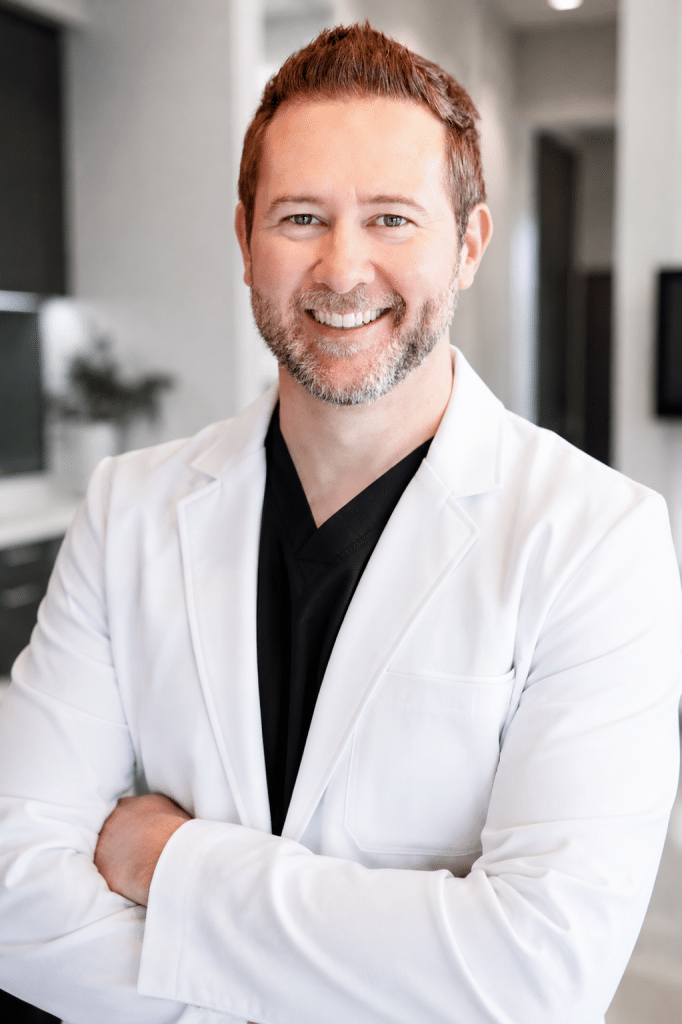 A smiling man with a beard and reddish-brown hair wears a white lab coat over a black scrub top.