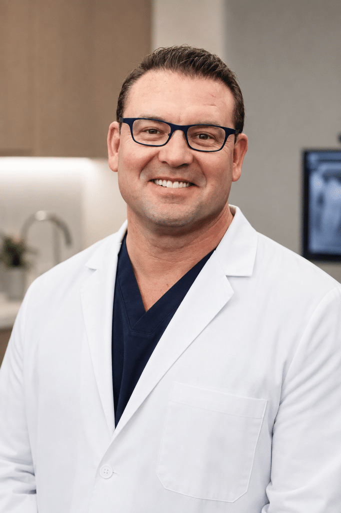 A smiling man in a white lab coat and blue scrubs wears glasses, looking directly at the viewer.