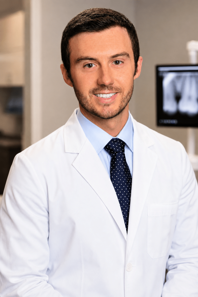 A smiling man with short dark hair and a beard wears a white lab coat and a blue polka-dot tie.